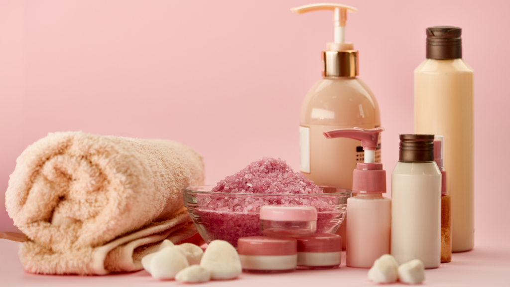 A collection of spa and skincare products displayed on a pink background, including towels, bottles of lotion, jars of cream, pink bath salts in a glass bowl, and small white decorative stones.