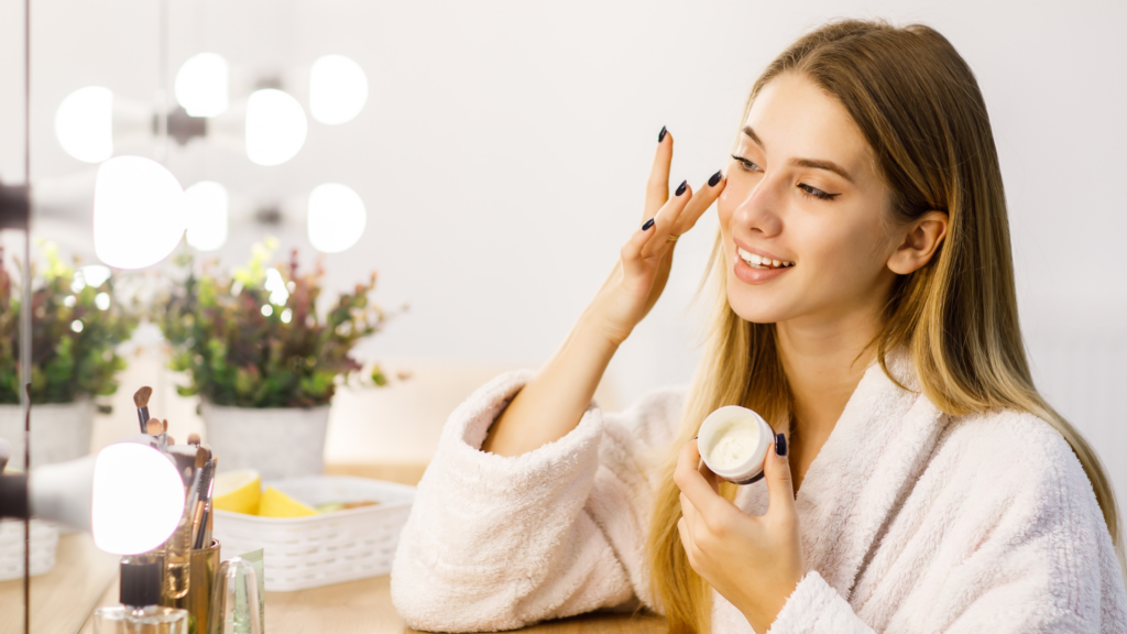 A young woman in a cozy bathrobe applies face cream while smiling in front of a mirror, surrounded by beauty products and soft lighting.