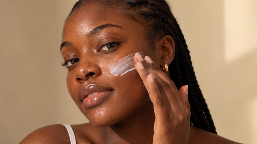 Woman with deep brown skin gently applying white sunscreen to her cheek against a warm beige background.
